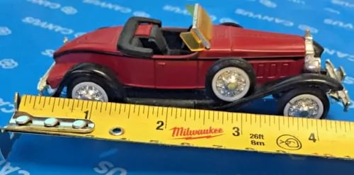 Red classic diecast model car with black roof and open hood, on a ruler scale