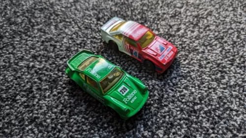 1/43 diecast model cars, green and red vintage-style coupes, angled on textured surface