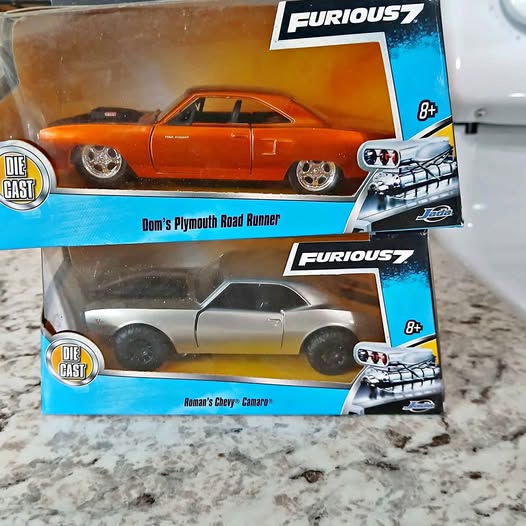 Two die-cast cars in retail boxes: orange Plymouth Road Runner and gray Camaro