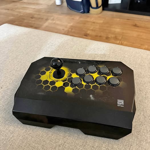 Black fighting stick controller with yellow hex pattern, joystick lever and round buttons