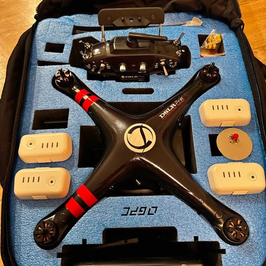 Black quadcopter drone with red accents and four beige battery modules in case.