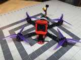 Freestyle FPV drone with red camera mount and purple propellers on black frame.