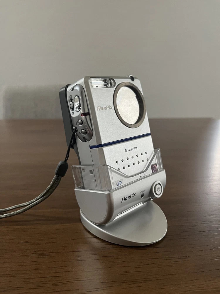 Silver compact digital camera with large lens and top screen, on a charging dock