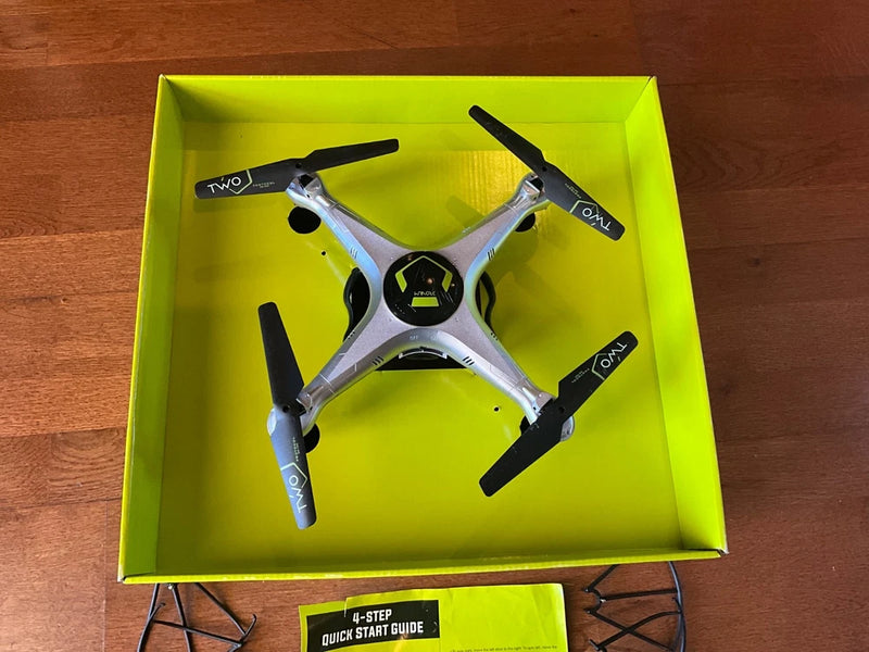 Silver and black quadcopter drone with four propellers, inside green packaging tray.