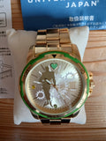 Gold-tone metal wristwatch with green bezel, gear dial, and stylized silhouette at 12