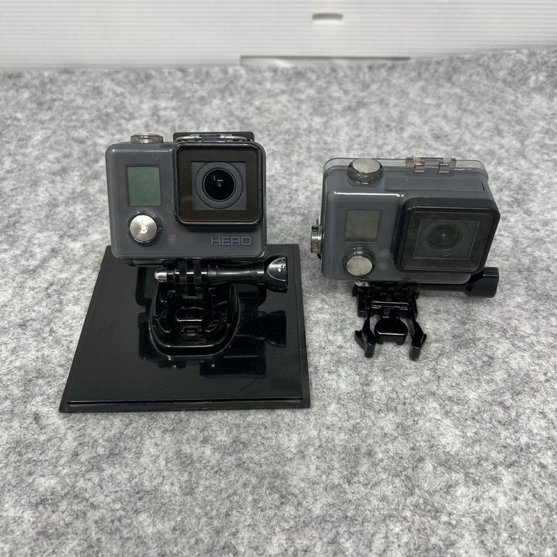 Two grey action cameras with LCD screens, waterproof housings, and mounts