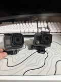 Two black action cameras in waterproof housings with mounts and doors, side-by-side