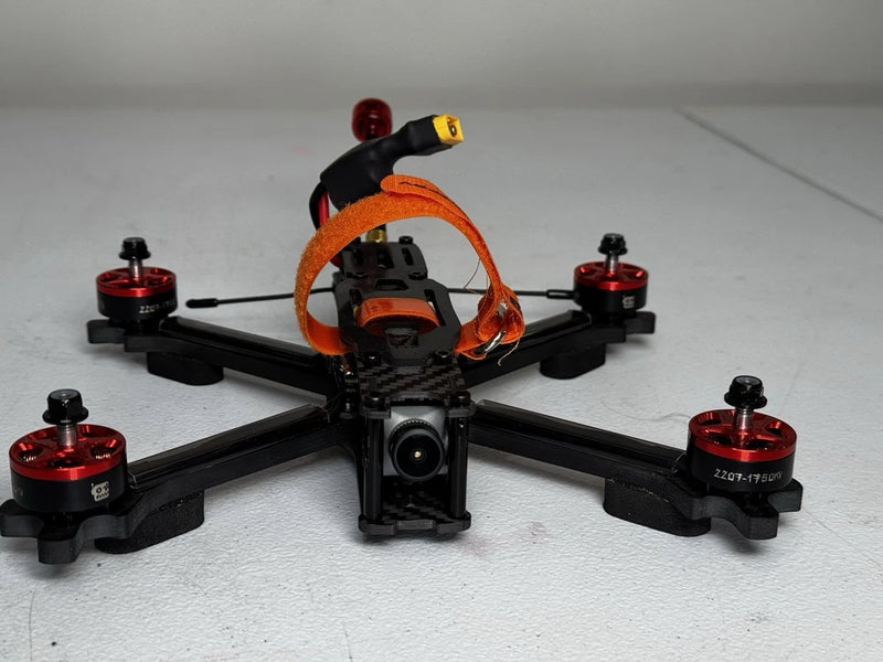 FPV quadcopter drone with black frame, orange prop guards, front camera lens