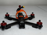 FPV quadcopter drone with black frame, orange prop guards, front camera lens