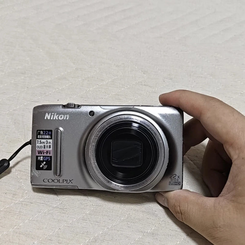 Silver Nikon COOLPIX S9500 digital camera with front lens and Nikon logo