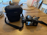 Digital camera with black FUJIFILM carrying case and wrist strap, showing rear LCD