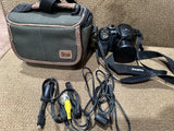 Black digital bridge camera with lens, neck strap, and cables; in fabric carrying case.