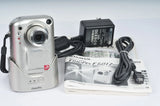 Silver compact digital camera with lens, wrist strap, and battery charger with cables
